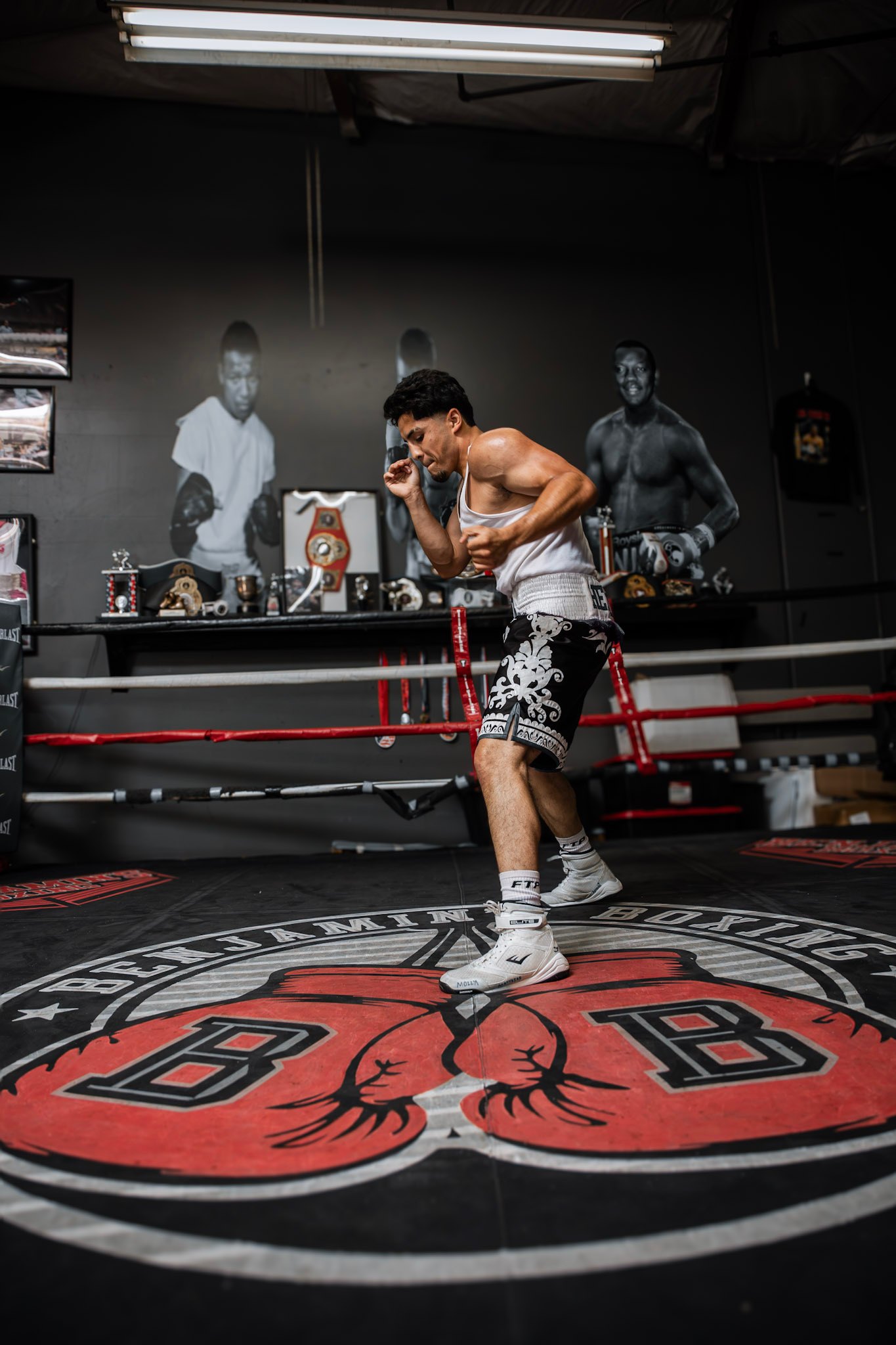 Juan Hernandez moving across the ring in fight trunks, portraits of past champions on the wall behind