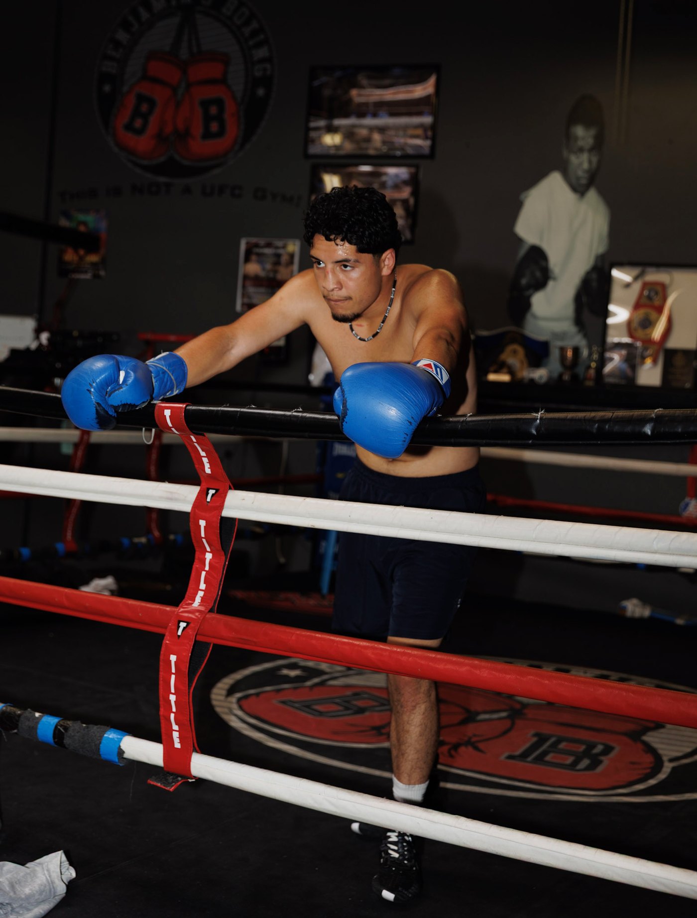 Juan Hernandez leaning on the top rope in blue gloves, the gym's wall of champions behind him